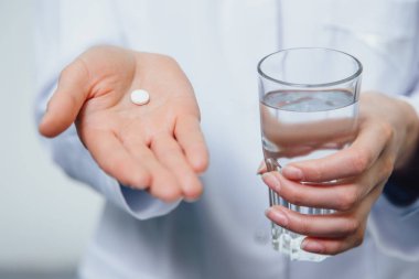 Genç kız doktor vitamin alır. Aynı zamanda, içinde beyaz bir elbise giymiş gri bir arka plan üzerinde duruyor. Bir bardak su ve bir hap elinde tutar.