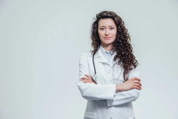 Mutlu genç gülümseyen kız doktor portresi. Beyaz bir elbise giymiş. Gri bir arka plan üzerinde çapraz eller ile eşit olarak ayakta.