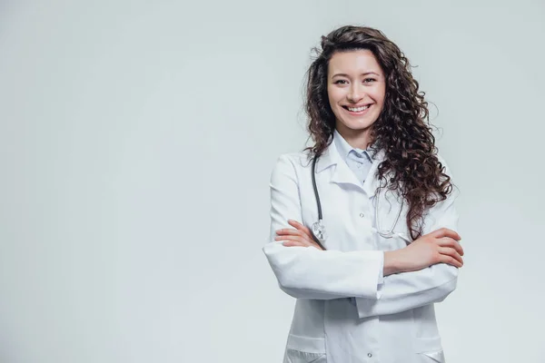 Mutlu genç gülümseyen kız doktor portresi. Beyaz bir elbise giymiş. Gri bir arka plan üzerinde çapraz eller ile eşit olarak ayakta.