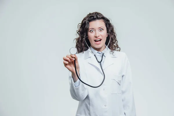 Uma jovem mulher caucasiana com um roupão de laboratório, a olhar para ti e a segurar o fonendoscópio fora de controlo. Retrato de uma mulher de cabelo muito escuro - um médico vestindo uniformes brancos. O — Fotografia de Stock