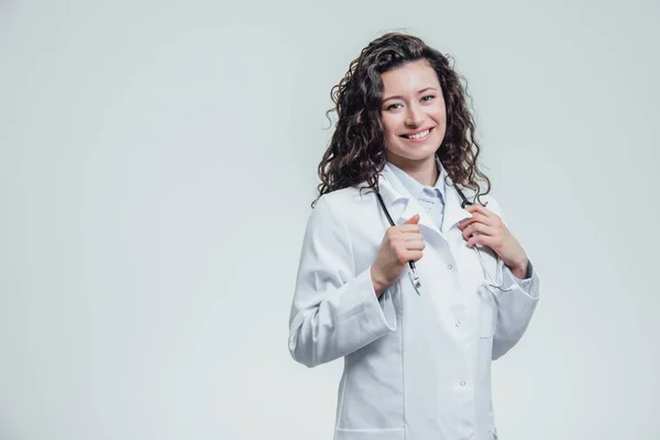 Uma jovem mulher caucasiana com um roupão de laboratório, a olhar para ti e a segurar o fonendoscópio fora de controlo. Retrato de uma mulher de cabelo muito escuro - um médico vestindo uniformes brancos. O — Fotografia de Stock