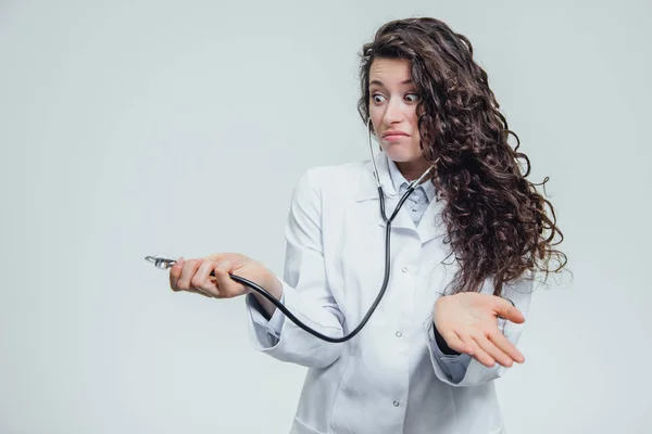 Uma jovem mulher séria em uma mulher caucasiana em um manto de laboratório, olhando para você e segurando o fonendoscópio em suas mãos tem um olhar surpreso. Retrato de uma mulher é cabelo muito escuro - um médico — Fotografia de Stock