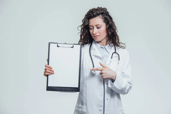 Молода жінка-лікар на сірому фоні. Під час цього він містить теку з документами. Показувати один з елементів . — стокове фото