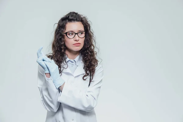 Schöne Krankenschwester mit schwarzen Haaren. trägt blaue uniformierte Handtaschen. in schwarzer Brille und Handschuhen vor grauem Hintergrund und blickt in die Kamera, ein Porträt. — Stockfoto