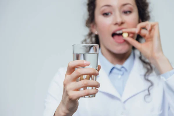 Una giovane dottoressa prende vitamine per le ossa. Allo stesso tempo, si erge su uno sfondo grigio vestito con una veste bianca. Tiene in mano un bicchiere d'acqua e una pillola bianca . — Foto Stock