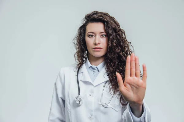 Uma jovem mulher bonita médico gestos pára. Provavelmente uma médica mostra um gesto parar sobre um fundo cinzento . — Fotografia de Stock