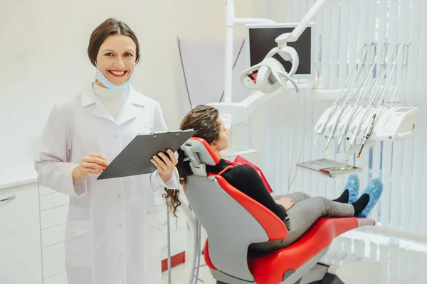 Un buen especialista. Retrato de una hermosa joven dentista vestida con bata. Volando contra el trasfondo de una clínica de odontología, sosteniendo una carpeta . — Foto de Stock