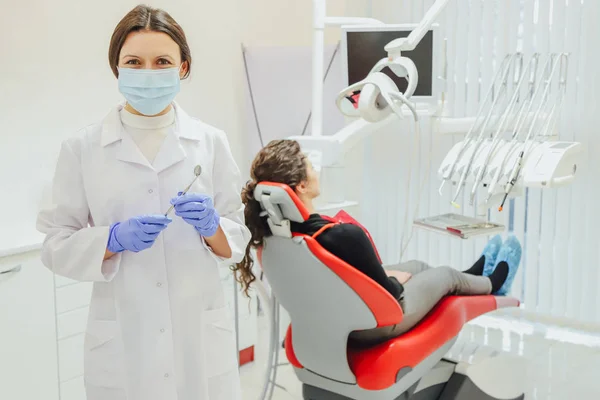 Un buen especialista. Retrato de una hermosa joven dentista vestida con bata y máscara. Contando la clínica de odontología, sosteniendo un espejo. Durante esto, el médico sonríe . — Foto de Stock