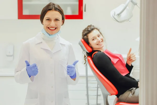 Un buen especialista. Retrato de una hermosa joven dentista vestida con bata y máscara. Dar a un dentista en el fondo de una clínica muestra una clase de gestos. Durante esto, el paciente también muestra una — Foto de Stock