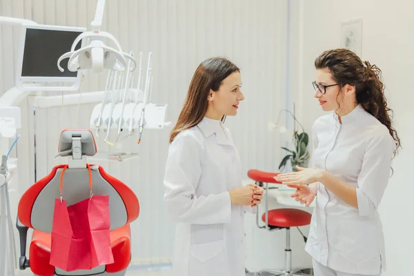 Dos doctores dentistas en el consultorio. Hermosas mujeres jóvenes. Ponte de pie y habla vestido con ropa blanca médica . — Foto de Stock