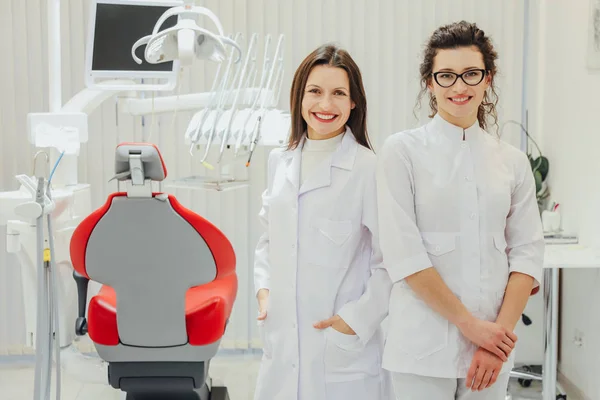 Dos doctores dentistas en el consultorio. Hermosas mujeres jóvenes. De pie con un toque el uno del otro. Mira la cámara. Vestido con ropa blanca médica. Durante esta sonrisa sincera . — Foto de Stock