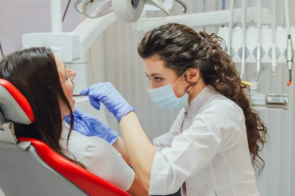 Mujer joven sentada en una silla dental para la cita de un médico. Durante este tiempo, ella estaba muy preocupada por el miedo, abrió sus grandes ojos. Un médico es una buena mujer con ropa médica, lo que hace un — Foto de Stock