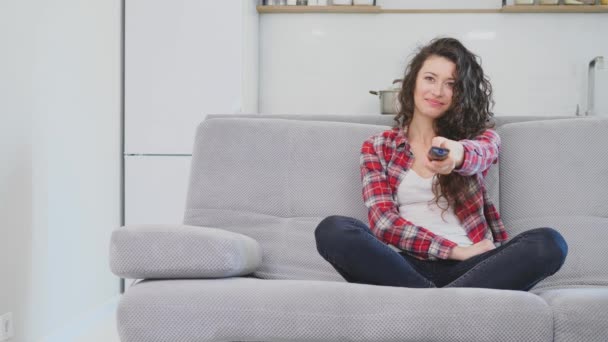 Une belle femme regarde la télévision et est assise sur le canapé et tient la télécommande dans sa main . — Video