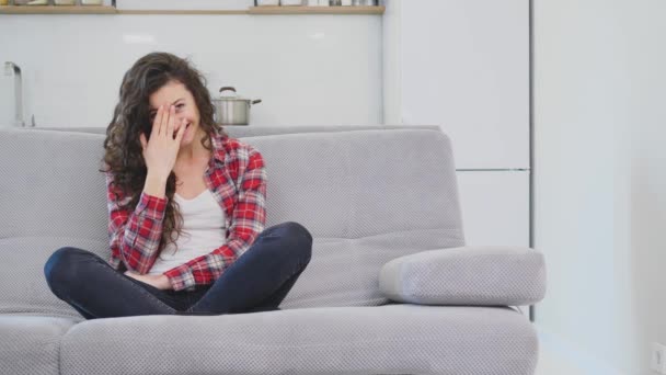 En vacker kvinna tittar på TV och sitter på soffan och håller fjärrkontrollen i handen. — Stockvideo