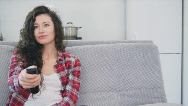 Uma mulher bonita assiste a TV e está sentada no sofá e segura o controle remoto em sua mão . — Vídeo de Stock