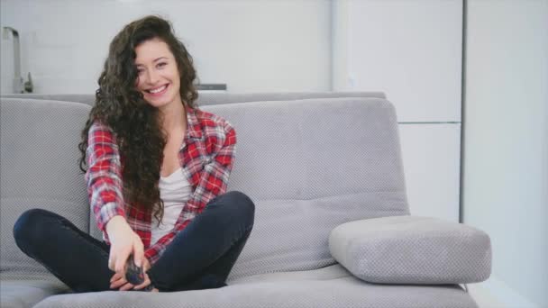 Eine schöne Frau schaut den Fernseher und sitzt auf der Couch und hält die Fernbedienung in der Hand. — Stockvideo