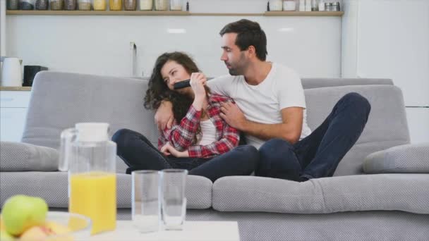 Όμορφο κορίτσι και ένας νεαρός άντρας κάθεται στον καναπέ και βλέπει τηλεόραση. Οι άνθρωποι χαλαρώνουν στον καναπέ. Νέος άντρας με γυαλιά και μπλε πουκάμισο. — Αρχείο Βίντεο