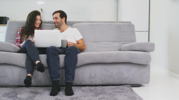 Молодая пара наслаждается утренним кофе с компьютером, сидя на диване дома вместе, счастливые мужчина и женщина весело смотрят на экран ноутбука . — стоковое видео