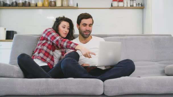 Sorridente giovane coppia godendo il caffè del mattino con un computer mentre seduti sul divano a casa insieme, felice uomo e donna guardando lo schermo del computer portatile divertimento . — Video Stock