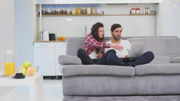 Χαμογελαστά Νεαρά ζευγάρια απολαμβάνουν τον πρωινό καφέ με έναν υπολογιστή, ενώ κάθονται στον καναπέ στο σπίτι μαζί, ευτυχισμένος άνθρωπος και γυναίκα κοιτάζοντας την οθόνη του φορητού υπολογιστή διασκέδαση. — Αρχείο Βίντεο