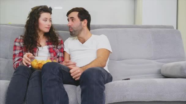 Leende fru och man sitter på soffan. Titta på den här TV: n. Håll dina marker med dina läppar och titta på kameran. — Stockvideo