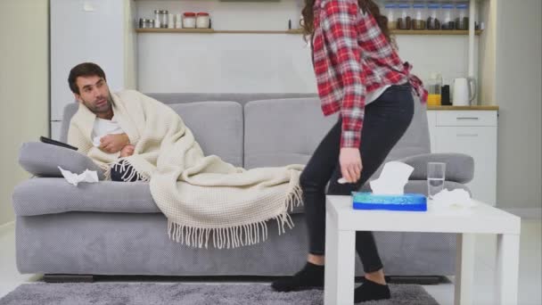 En man med en förkylning sitter på en soffa i en filt filt. Kontrol lera temperaturen. Hustrun sitter bredvid henne och hjälper till. Man hemma. Sjukt ung man grå soffa i rummet. Olycklig kille. Koncept — Stockvideo
