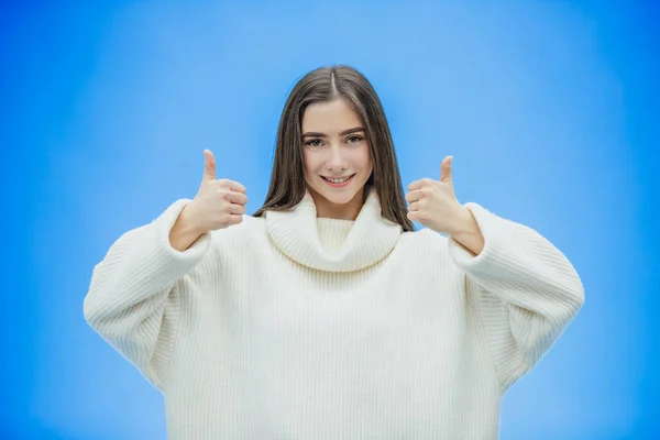 Menina bonita em um fundo azul. A menina mostra um telefonema com um gesto. Vestido com uma camisola branca quente. Tem cabelo preto longo . — Fotografia de Stock