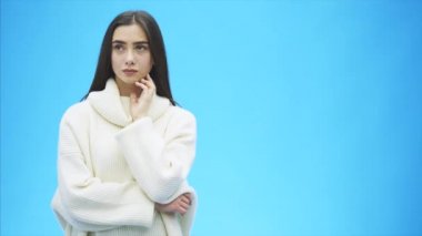 Genç kız mavi bir arka plan üzerinde duruyor. Bu sırasında ciddi, çene altında ellerini koyarak. Beyaz bir kazak Dressed düşünme sonra.