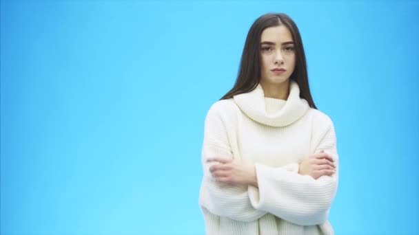 Mladá dívka stojí na modrém pozadí. Během této vážné, položí ruku na rameno. Oblečený v bílém svetru. Podívejte se do kamery. — Stock video