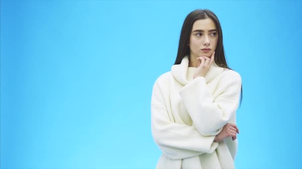 Ung flicka står på en blå bakgrund. Under denna allvarliga, sätta händerna under hakan. Efter att ha funderat Dressed i en vit tröja. — Stockvideo