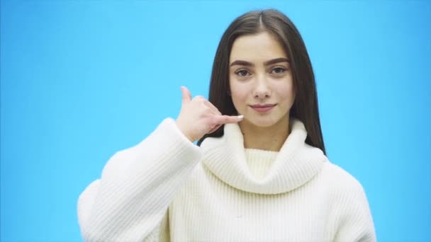 Krásná mladá dívka na modrém pozadí. Dívka ukazuje telefonní hovor s gestem. Oblečený v bílém svetru. — Stock video