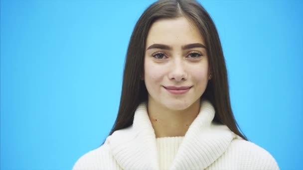 Schönes junges Mädchen, das auf blauem Hintergrund steht. Während dieser Zeit trägt sie einen weißen Pullover. mit langen schwarzen Haaren. — Stockvideo