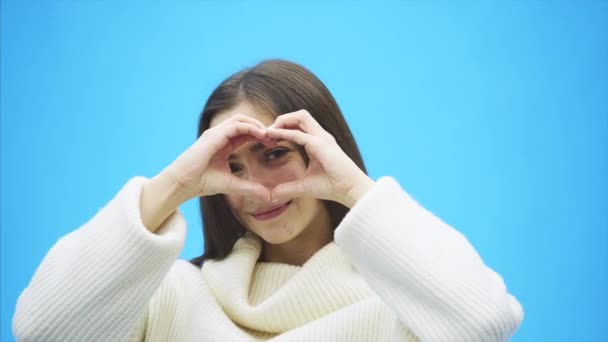 Portrait of a happy young woman wearing a white warm sweater. During this time, a hand gesture of heart shows and feels love. Isolated on a blue background. Happy and smiling. — Stock Video