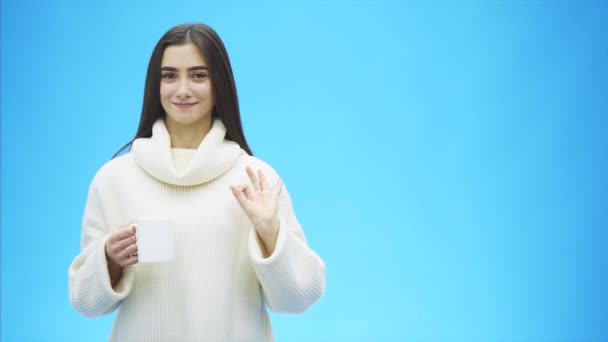Curiosamente, una mujer joven en un suéter blanco de punto. Lleva una taza blanca de café o té en las manos. Aislado sobre el fondo de una pared azul, retrato de estudio. Concepto de una forma de vida de — Vídeos de Stock
