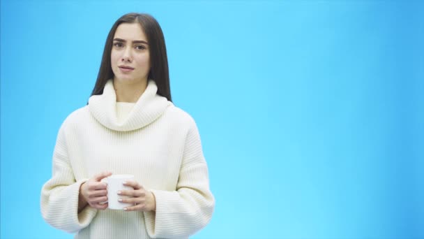 Curiosamente, uma jovem mulher em uma camisola branca de malha. Tem uma chávena branca de café ou chá nas mãos. Isolado contra o fundo de uma parede azul, retrato de estúdio. Conceito de um modo de vida de — Vídeo de Stock