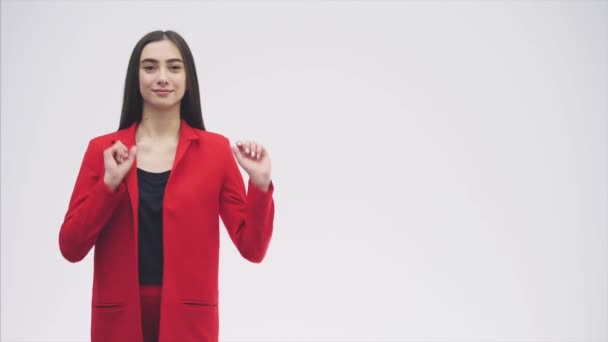 Retrato de una hermosa joven morena. Mira a la cámara. Durante esto, levantó las manos hacia arriba apuntando con el dedo hacia un lado. Vestido con una chaqueta roja. Fondo gris . — Vídeos de Stock