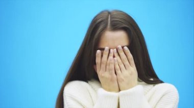 Güzel genç mavi bir arka plan üzerinde duran kız. Bu süre içinde o beyaz bir sıcak Kazak giymiş. Gülümser ve bunun ardından ellerini kapatarak yüz açar.