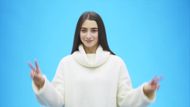 Young beautiful brunette woman on an isolated background. He raised his hands and showed a gesture with two fingers. Smiling confidently and happily. On a blue background. Dressed in a white sweater. — Stock Video