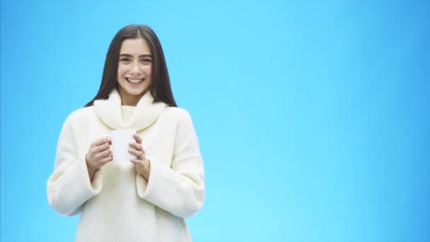 Curiosamente, una mujer joven en un suéter blanco de punto. Lleva una taza blanca de café o té en las manos. Aislado sobre el fondo de una pared azul, retrato de estudio. Concepto de una forma de vida de — Vídeo de stock