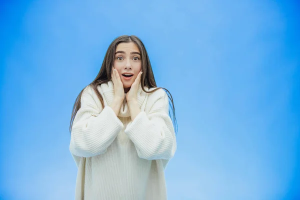 Μια νεαρή γυναίκα, φορώντας ένα πουλόβερ του χειμώνα σε ένα απομονωμένο γαλάζιο φόντο, χαμογελώντας στην αγάπη, υψώνοντας το χέρι από έκπληξη βάζει στο πηγούνι. Ωραίο κορίτσι. — Φωτογραφία Αρχείου