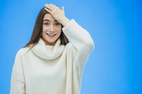 Una mujer joven con un suéter de invierno sobre un fondo azul aislado, sin sonreír, levantando la mano del dolor las pone en la frente . —  Fotos de Stock