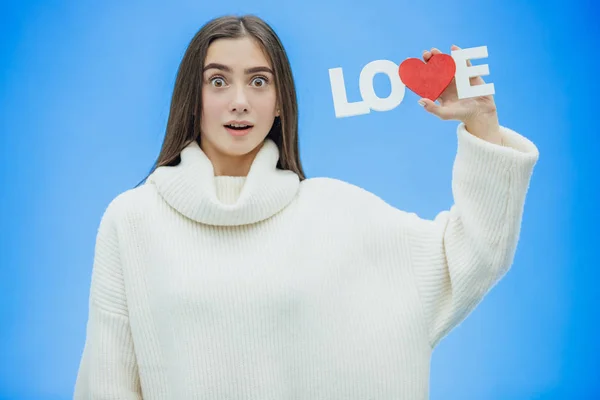 Menina bonita nova vestida com roupas brancas. Durante este tempo ele está em um fundo azul. Mantém uma palavra de amor nas mãos . — Fotografia de Stock