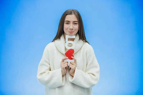 Menina bonita nova vestida com roupas brancas. Durante este tempo ele está em um fundo azul. Mantém uma palavra de amor nas mãos . — Fotografia de Stock