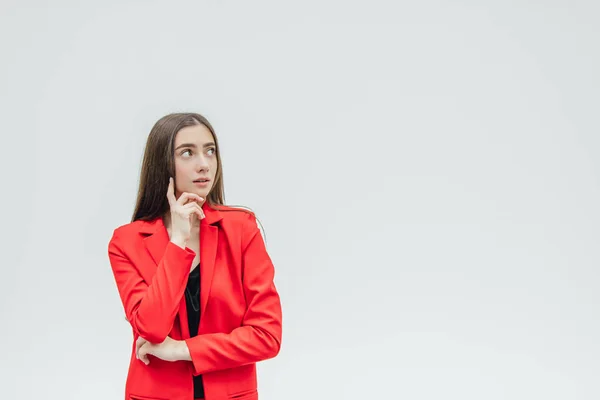 The young pretty girl is on a gray background. During this time, he put his hand under the chin. Serious Dressed in a red jacket. — Stock Photo, Image