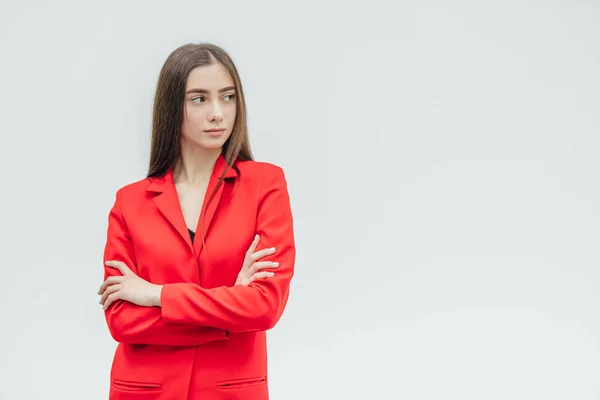 Young pretty, serious girl on a gray background. Standing with a hand on hand. Dressed in a red jacket. With black long hair. — Stock Photo, Image