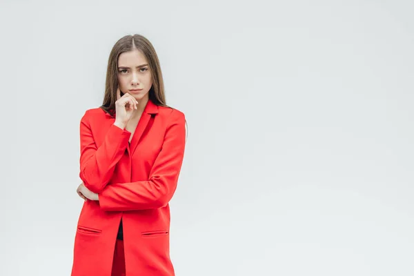 Young pretty, serious girl on a gray background. Standing with a hand on hand. Dressed in a red jacket. With black long hair. — Stock Photo, Image