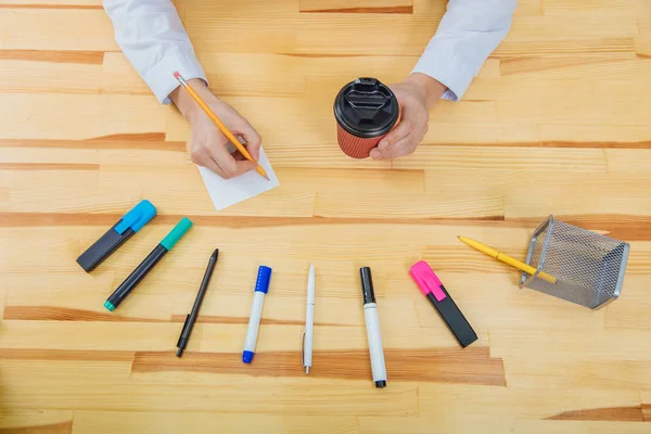 Mesa de escritório com acessórios: uma folha branca de papel, um marcador azul-rosa, um copo de café, uma caneta, um lápis e mãos de mulheres . — Fotografia de Stock