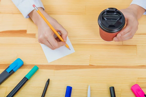 Mesa de escritório com acessórios: uma folha branca de papel, um marcador azul-rosa, um copo de café, uma caneta, um lápis e mãos de mulheres. Durante esta mão segura um copo de café, outro lápis escreve sobre — Fotografia de Stock