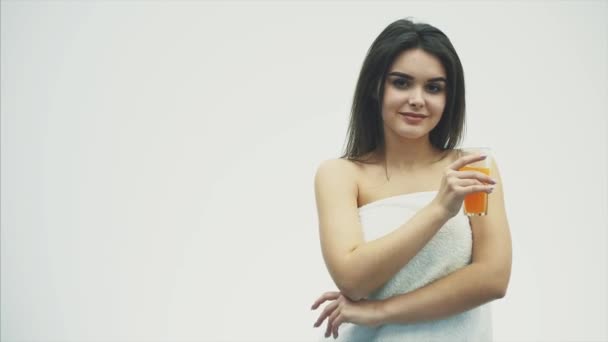 A jovem bela mulher bebe suco de laranja doce de um copo transparente com um sorriso, sobre um fundo branco. Com cabelo longo preto bonito . — Vídeo de Stock