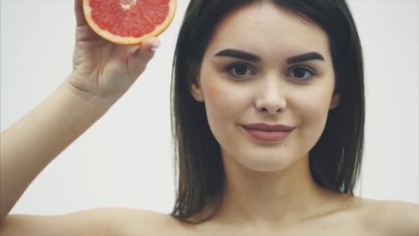 Portrait of a beautiful young woman standing looking at the camera. Takes care of your body using fruit and making masks. Holding a half orange fruit on her shoulders with long black hair, isolated on — Stock Video